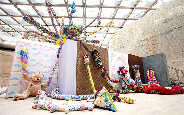 A large-scale, colorful installation filled with crocheted creatures and objects. The installation includes a giant, crocheted spider, a colorful teddy bear, and various other creatures and forms.